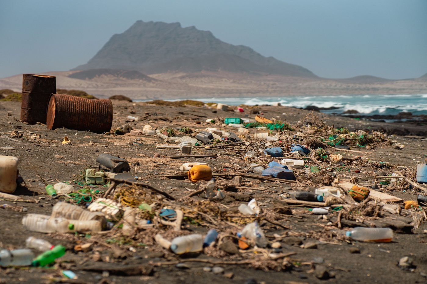 Bouteilles, contenants de plastique et filets de pêche échoués sur une plage de sable noir près de barils rouillés.