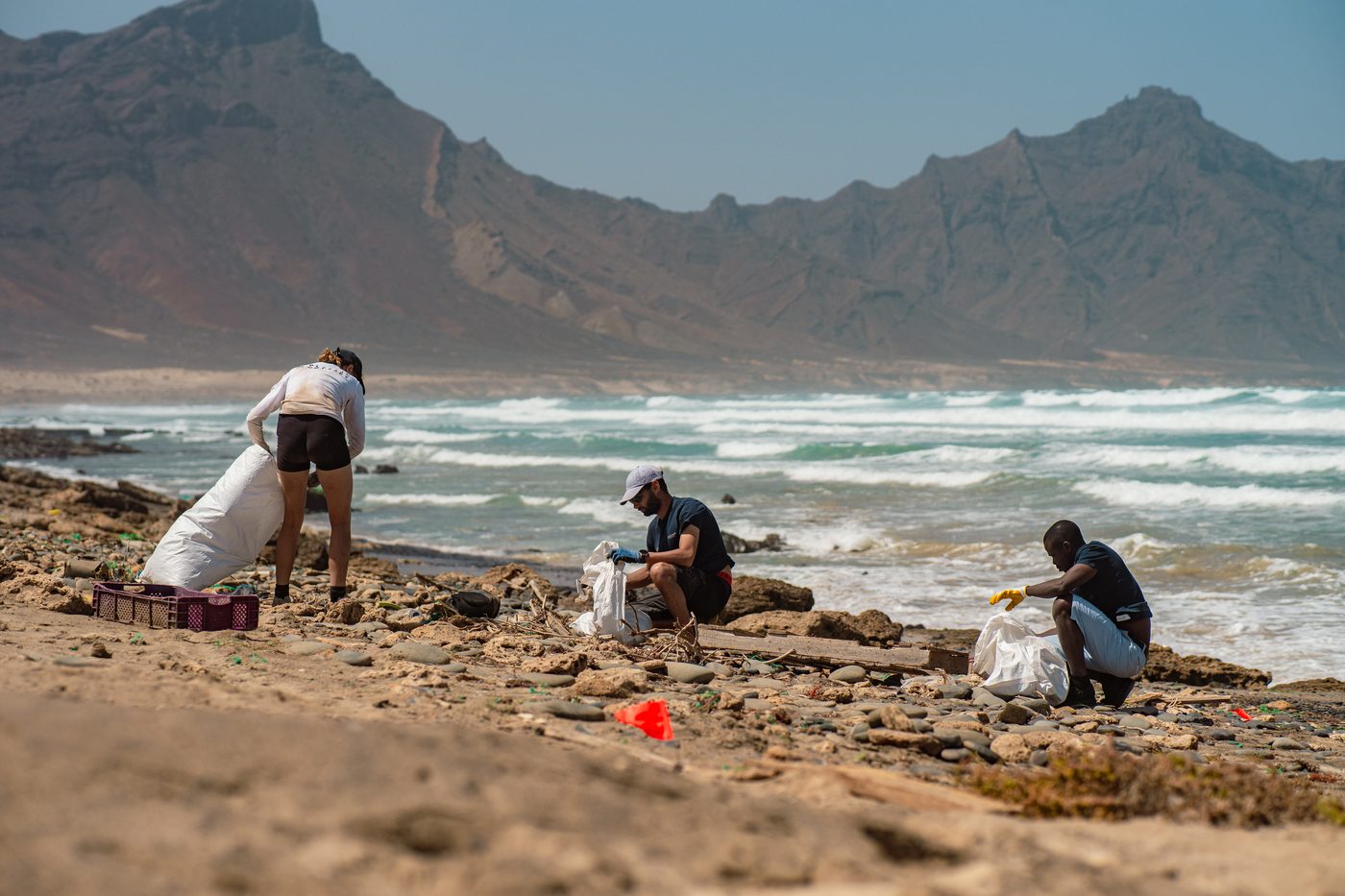 Crew members fill bags with plastic debris on a rocky shoreline below volcanic peaks.