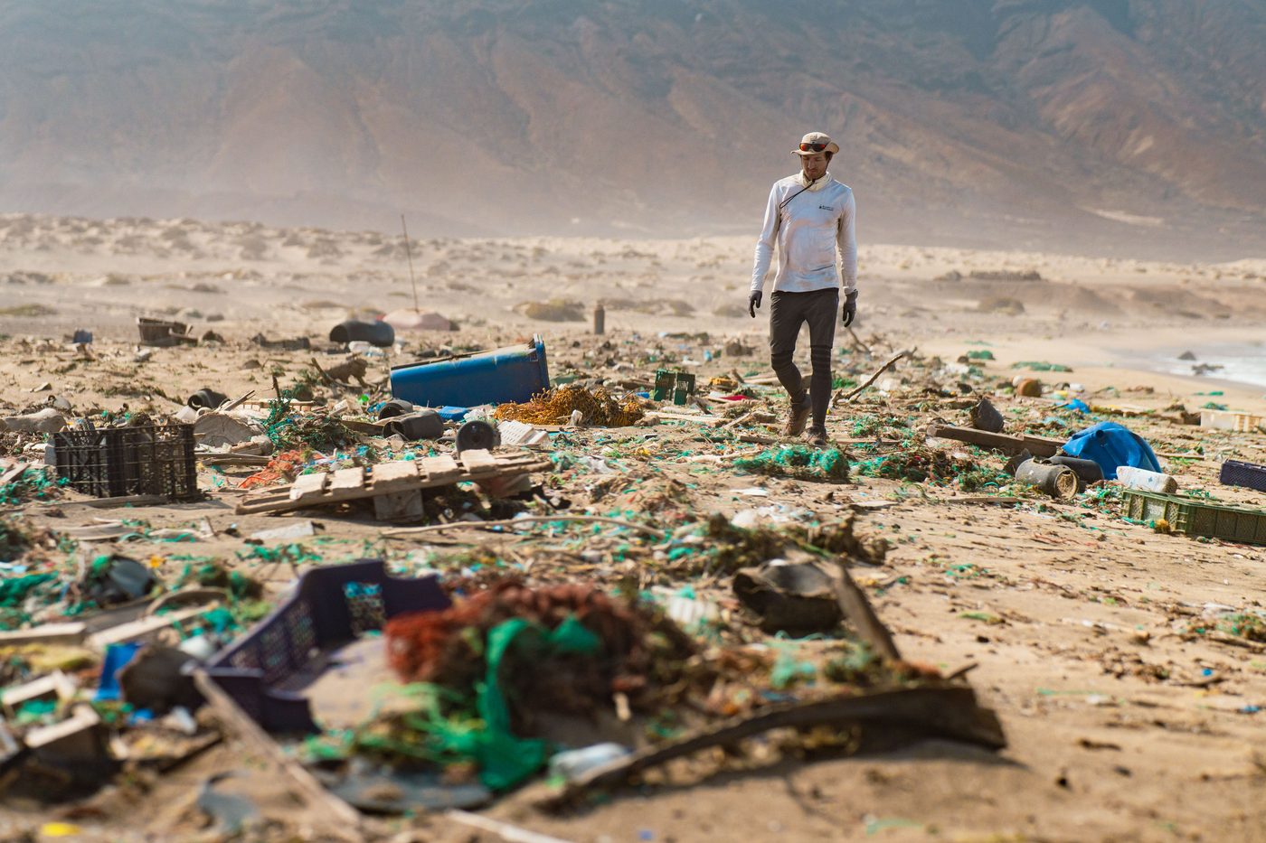 A crew member surveys a shoreline strewn with plastic crates, nets and drums.